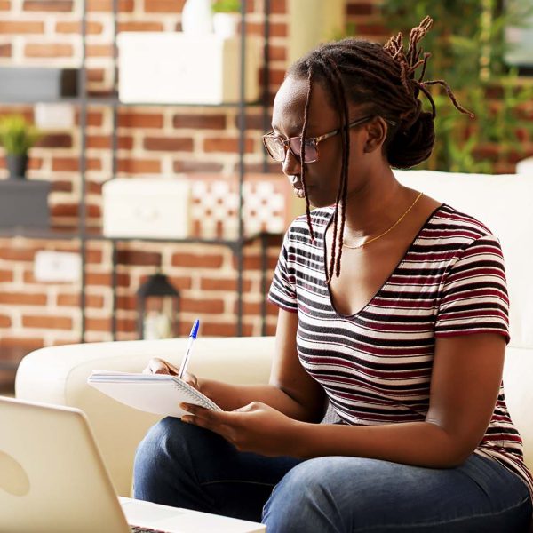 African american student taking notes at online class lesson, using notebook to write business information. Female freelancer browsing internet on laptop and attending school webinar.